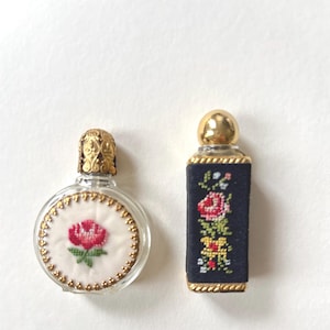 Könnte beinhalten: Zwei antike Parfümflaschen. Eine ist rund mit einer roten Rosenstickerei auf weißem Stoff und einem goldenen Filigranaufsatz. Die andere ist rechteckig mit einem schwarzen Stoff, der mit einem floralen Design bestickt ist, und einer goldenen Kappe.