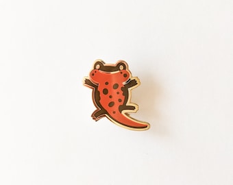 May include: A gold-tone enamel pin featuring a red and black salamander with a smiling face. The salamander is depicted with black spots and a long tail.