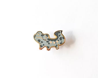 May include: A gold-toned enamel pin featuring a gray and white spotted salamander with a light pink belly. The salamander is facing right and has a slightly curved tail.