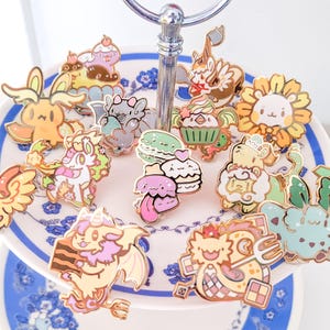 May include: A collection of colorful enamel pins featuring various cartoon creatures, animals, and food items like cupcakes and macarons. The pins are arranged on a tiered white serving tray with blue floral patterns and a silver metal handle.