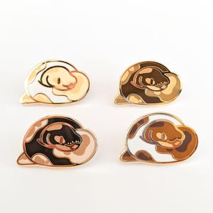 May include: Four enamel pins shaped like coiled snakes, each featuring a different color palette of white, tan, brown, and black. The pins have gold-toned metal outlines and depict a snake curled into a rounded, cloud-like shape.