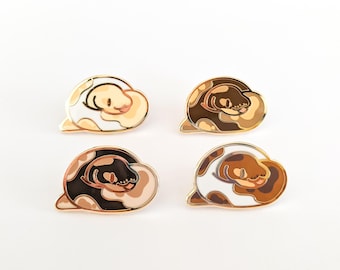 May include: Four enamel pins shaped like coiled snakes, each featuring a different color palette of white, tan, brown, and black. The pins have gold-toned metal outlines and depict a snake curled into a rounded, cloud-like shape.