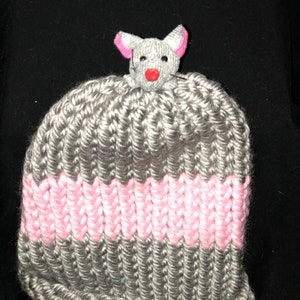 May include: A gray and pink knitted hat with a gray mouse-shaped topper. The mouse has pink ears and a red nose.