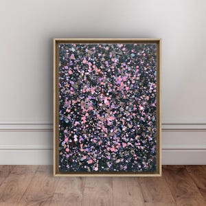 May include: Abstract art piece in a wooden frame. The artwork features a dark background covered in a pattern of pink, purple, and silver paint splatters. The frame is a light brown color, and the artwork is displayed against a white wall.