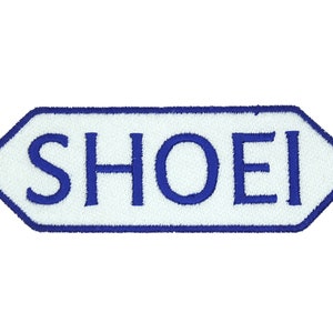 Peut inclure: Un écusson hexagonal blanc avec une bordure bleue et le mot "SHOEI" brodé en bleu.