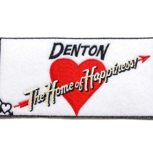Puede incluir: Parche blanco bordado con contorno negro. El parche presenta un corazón rojo con una flecha que lo atraviesa y el texto "Denton The Home of Happiness!" en una fuente cursiva blanca.