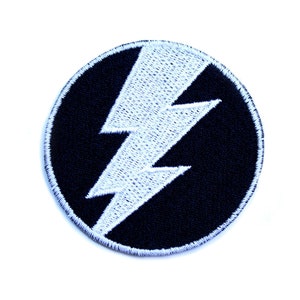 Puede incluir: Un parche bordado en blanco y negro con un diseño de rayo. El parche es circular y tiene un contorno blanco.