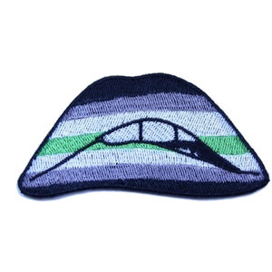 May include: An embroidered patch in the shape of a pair of lips with the colors of the genderqueer pride flag. The flag is made up of stripes of gray, green, and white.