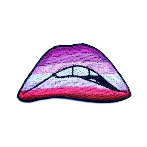 Puede incluir: Un parche bordado en forma de labios, con rayas rosas, blancas y azules. Los labios están ligeramente entreabiertos, mostrando una fila de dientes blancos.