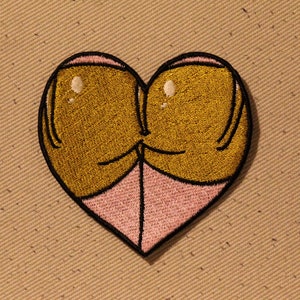 May include: A heart-shaped embroidered patch with two golden-brown figures hugging each other. The figures are outlined in black and the heart is filled with a light pink color.