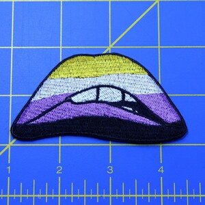 Puede incluir: Un parche bordado en forma de labios con un esquema de color amarillo, blanco, morado y negro. Los colores representan la bandera del orgullo no binario.