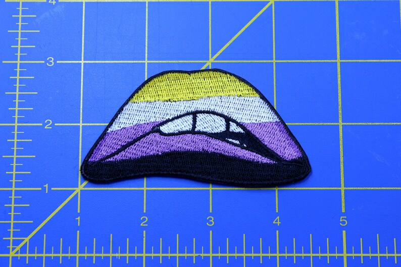 Puede incluir: Un parche bordado en forma de labios con un esquema de color amarillo, blanco, morado y negro. Los colores representan la bandera del orgullo no binario.