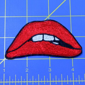 Może przedstawiać: Czerwone haftowane usta z białymi zębami i czarnym obramowaniem. Usta są lekko rozchylone, odsłaniając zęby. Hafty są szczeg&oacute;łowe i realistyczne.