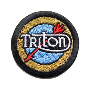 May include: A black and gold embroidered patch with the word "Triton" in white lettering and a red arrow pointing to the right.