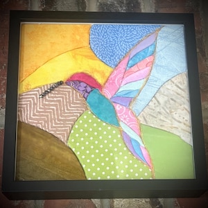 May include: A square framed fabric collage depicting a hummingbird in flight. The bird is composed of various patterned textiles in shades of pink, blue, and teal, while the background features yellow, green, and light blue fabric sections.