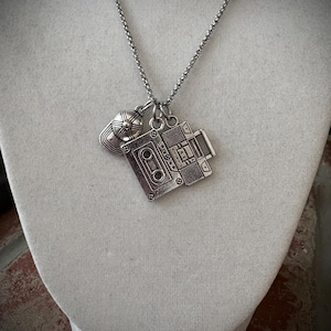 May include: Silver-toned necklace featuring three charms: a baseball cap, a cassette tape, and a video camera. The charms are detailed with etched designs and hang from a delicate chain. The necklace is displayed on a white mannequin.