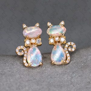 May include: A pair of gold-toned cat-shaped earrings. Each earring features an opal head and a pear-shaped opal body. The earrings are embellished with small, sparkling stones and have curled tails.