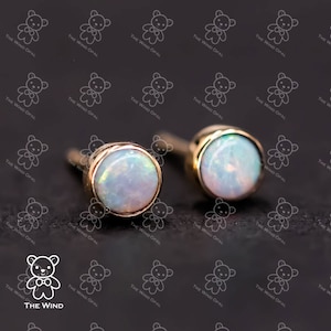 May include: A pair of gold-colored stud earrings with iridescent opal stones. The round opals are set in a bezel setting. The earrings are displayed on a dark surface with a repeating pattern of teddy bear logos and the text "THE WIND OPAL".