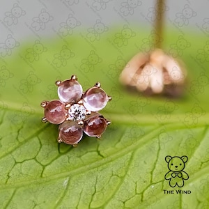 Puede incluir: Un pendiente de botón en forma de flor, con un diamante central y cinco gemas de color rosa-púrpura. El pendiente es de oro y descansa sobre una hoja verde. El logotipo de The Wind Opal está en la esquina inferior derecha.