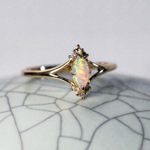 May include: A gold ring featuring a central marquise-cut opal gemstone with a vibrant play-of-color. The setting includes small, clear accent stones arranged in a cluster above and below the opal, set on a split-shank gold band.