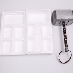 Pu&ograve; includere: Un martello color argento con un manico in pelle marrone e un inserto di imballaggio in schiuma bianca. Il martello ha una superficie testurizzata e un design decorativo sulla testa.