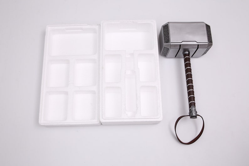 Pu&ograve; includere: Un martello color argento con un manico in pelle marrone e un inserto di imballaggio in schiuma bianca. Il martello ha una superficie testurizzata e un design decorativo sulla testa.