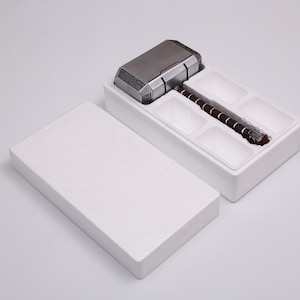 Pu&ograve; includere: Una scatola bianca con un interno in schiuma, aperta per rivelare un martello di metallo argentato e marrone con un manico. Il martello &egrave; una replica del martello di Thor, Mjolnir, dei fumetti e dei film Marvel.