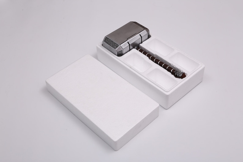 Pu&ograve; includere: Una scatola bianca con un interno in schiuma, aperta per rivelare un martello di metallo argentato e marrone con un manico. Il martello &egrave; una replica del martello di Thor, Mjolnir, dei fumetti e dei film Marvel.