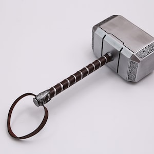 Pu&ograve; includere: Un martello color argento con un manico in pelle marrone e un cinturino. Il martello presenta dettagli intricati e una superficie testurizzata.