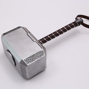 Pu&ograve; includere: Un martello color argento con un manico in pelle marrone e un cinturino decorativo. Il martello ha un design dettagliato sulla testa e un'incisione circolare sul lato.