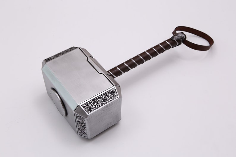 Pu&ograve; includere: Un martello color argento con un manico in pelle marrone e un cinturino decorativo. Il martello ha un design dettagliato sulla testa e un'incisione circolare sul lato.