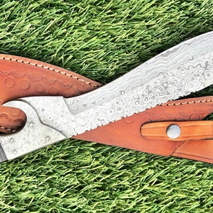 May include: A silver-colored knife with a curved blade and a brown leather sheath. The knife has a decorative pattern on the blade and a black handle.
