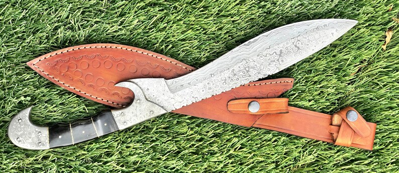 May include: A silver-colored knife with a curved blade and a brown leather sheath. The knife has a decorative pattern on the blade and a black handle.