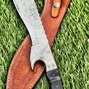 May include: A silver knife with a patterned blade and a black handle. The knife is in a brown leather sheath with a decorative stitching pattern.