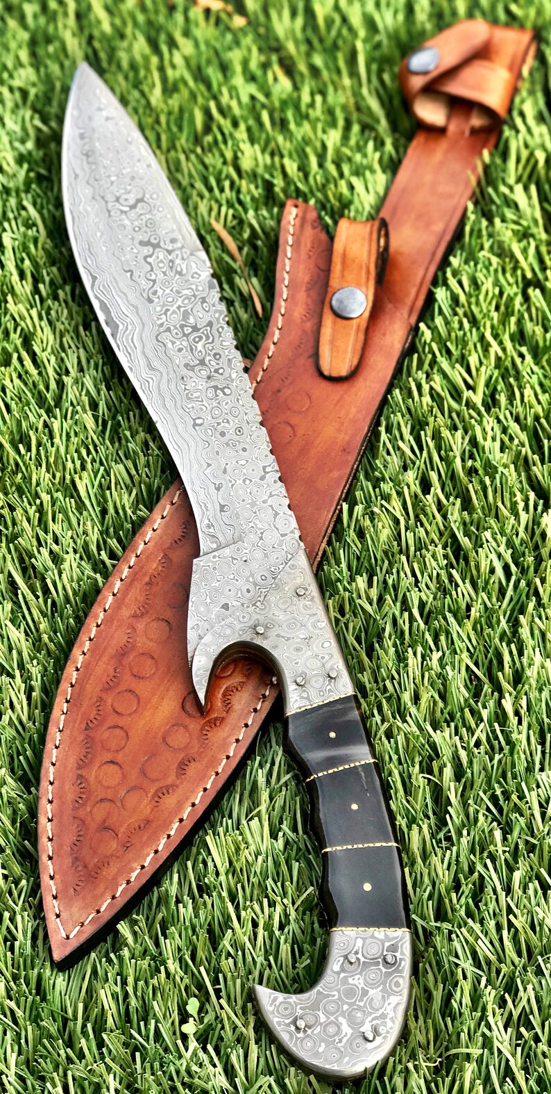 May include: A silver knife with a patterned blade and a black handle. The knife is in a brown leather sheath with a decorative stitching pattern.