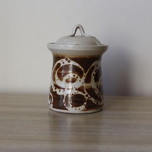 May include: A white ceramic jar with a brown swirl design and a lid with a handle. The jar is approximately 15 cm tall.