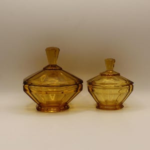 May include: Two amber-coloured glass bowls with matching lids, each featuring a faceted, geometric design and a pointed finial handle. The bowls have a wide, hexagonal base and a tapered shape, showcasing a translucent, warm golden hue throughout.
