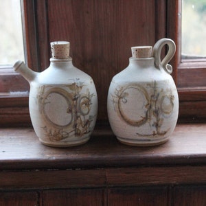 May include: Two ceramic jugs with cork stoppers and a hand-painted design. The jugs are white with brown and beige accents. The jugs are sitting on a wooden windowsill.