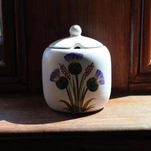 May include: A white ceramic sugar bowl with a rounded body and a matching lid featuring a small knob handle. The front of the bowl is decorated with a painted illustration of three purple thistle flowers with green stems and leaves.