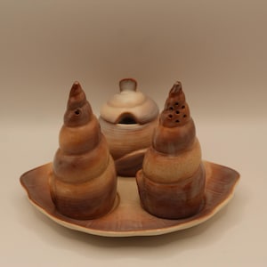 May include: A ceramic salt and pepper shaker set and a small lidded container. The set is brown and tan, with a seashell design. The set sits on a matching leaf-shaped tray.