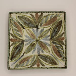May include: Square ceramic plate with a floral design. The plate features a central flower pattern in shades of blue and brown, surrounded by stylized leaves in green and brown. The border is a muted green.
