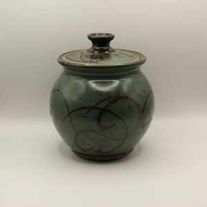 May include: A green ceramic jar with a lid. The jar is round and adorned with dark brown swirling patterns. The lid is a lighter green with similar brown designs and a dark brown knob. A decorative storage container.