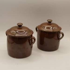 May include: Two matching brown ceramic pots with lids. Each pot has a rounded body, small handles, and a lid with a small knob. The pots are a glossy, dark brown colour, and are likely for cooking or storage.