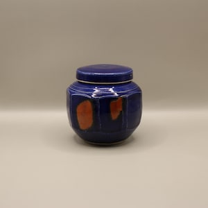 May include: A small, blue ceramic jar with a lid. The jar has a geometric design and orange accents.