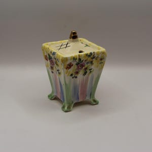 May include: A vintage ceramic sugar bowl with a square shape and a lid. The bowl is decorated with colourful floral patterns and vertical stripes in pastel colours. The lid has a gold-coloured handle and a crosshatch design.