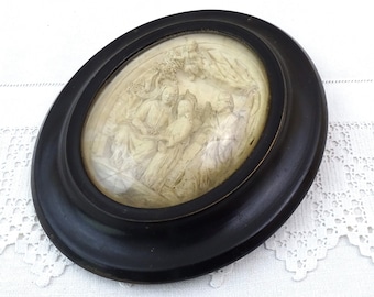 May include: An oval-shaped framed artwork with a black wooden frame and a glass cover. The artwork depicts a white relief sculpture of a religious scene with three figures.