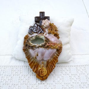 May include: A ceramic wall hanging in the shape of a heart, featuring a cross at the top and intricate details. The heart is a mix of colors, including brown, gold, and pink, with a small opening in the center. The piece is set against a white pillow.