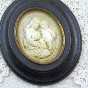 May include: A framed oval cameo with a white relief sculpture of the Virgin Mary holding a baby Jesus. The frame is black with a gold inner border.