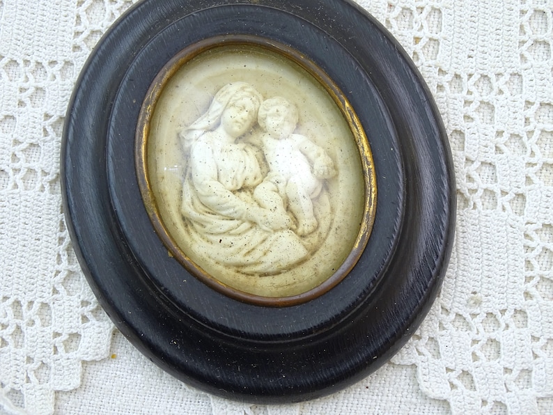 May include: A framed oval cameo with a white relief sculpture of the Virgin Mary holding a baby Jesus. The frame is black with a gold inner border.