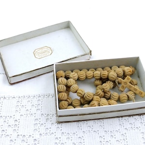 May include: A vintage rosary with carved wooden beads and a cross, presented in a white box with a lid. The beads have a detailed, ribbed design. The box has a label on the inside of the lid.